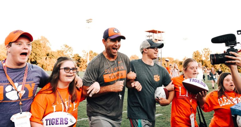 This photo shows Clemson Head Football Coach Dabo Swinney leading a group of young people with intellectual disabilities onto the field before a football game.