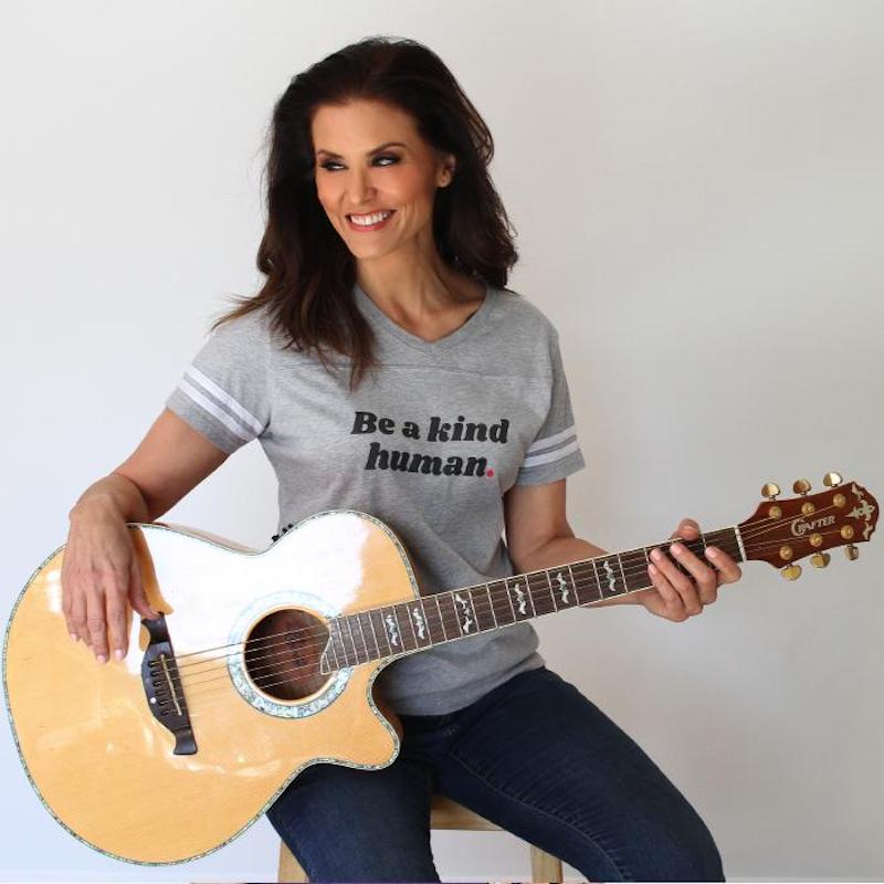 this photo shows former Miss USA Lu Parker wearing a t-shirt from Be Kind & Co. and holding an acoustic guitar