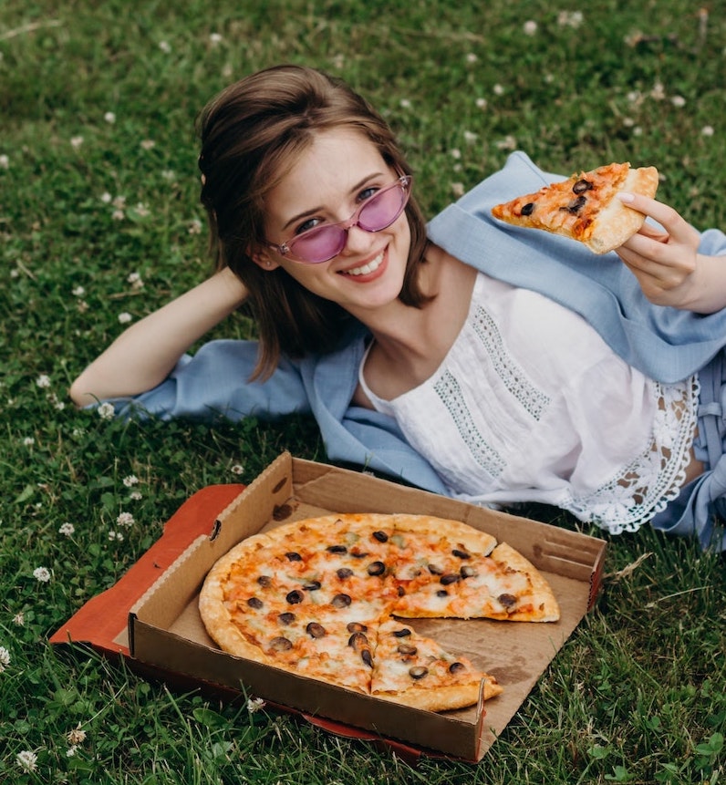 this photo shows a young woman eating pepperoni pizza from a recyclable pizza box that has a visible grease stain on the bottom.