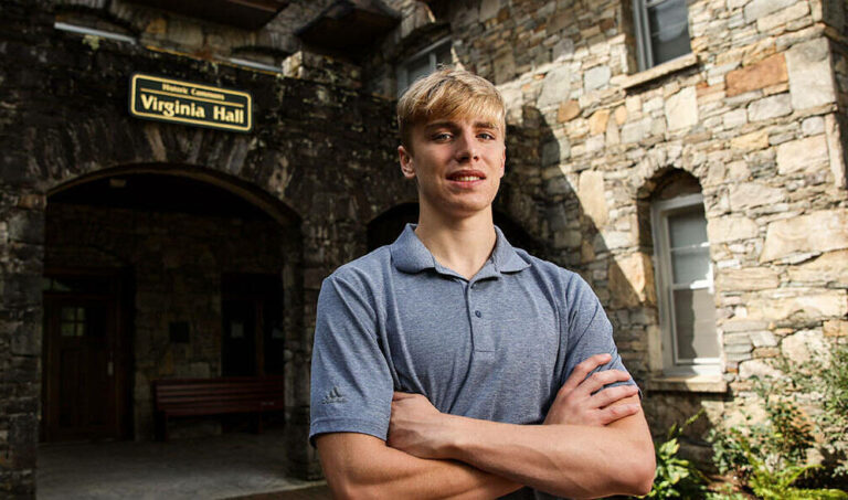 this photo shows Matthew Knafelz, a young man with blonde hair and wearing a blue shirt, posing with arms crossed in front of Virginia Hall at Lees-McRae College.